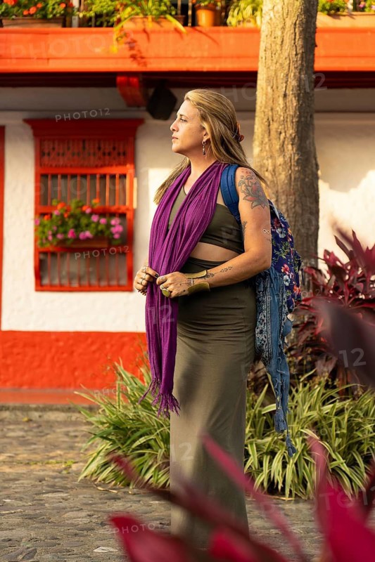A confident woman wearing a purple shawl, tattoos and a backpack explores a bustling Caribbean street surrounded by tropical vegetation and vibrant red and white architecture.