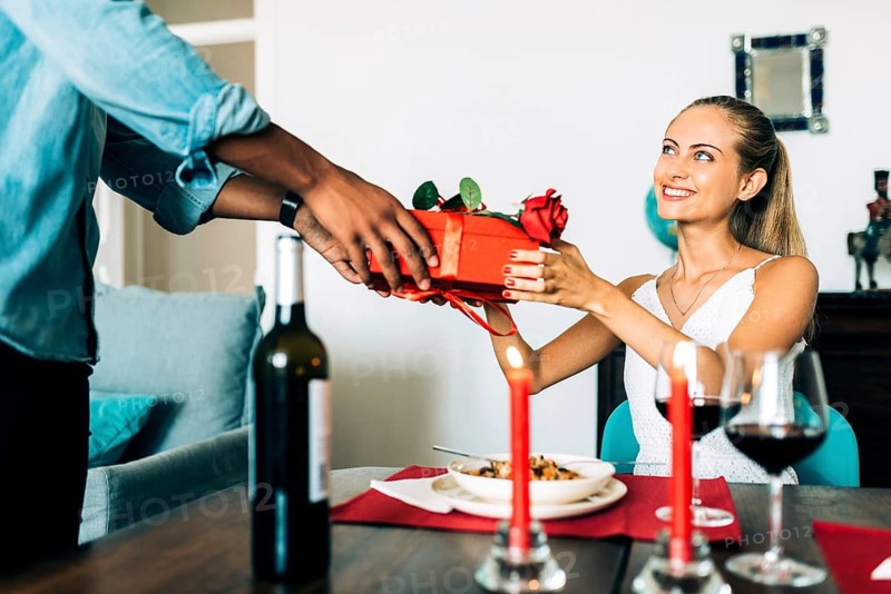 A couple shares a gift in a cozy setting with a candlelit dinner. The woman, smiling, receives a red-wrapped present on a table set with wine and food, creating a romantic atmosphere