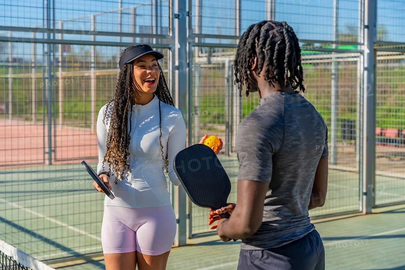Two diverse young pickleball players smiling and chatting while holding paddles and a ball on an outdoor court, enjoying friendly sport, fitness and warm weather fun