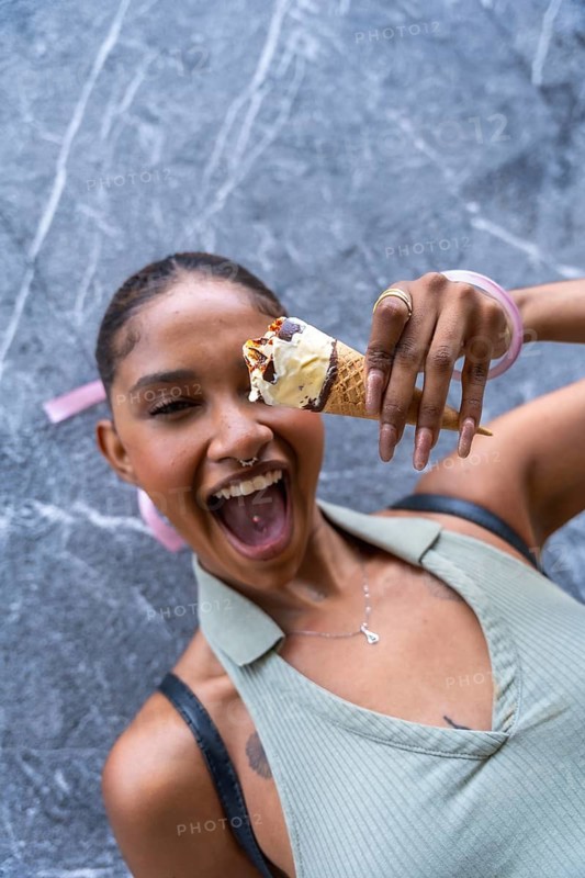 Joyful woman with a wide open mouth laughs while holding a delicious vanilla ice cream cone with chocolate syrup and toppings against a marble wall, expressing happiness and summer leisure
