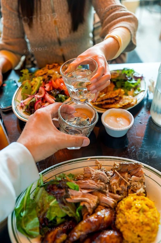 Two people are clinking small glasses together over a table filled with various dishes, including meat, rice, and salads, sharing a celebratory moment during a meal
