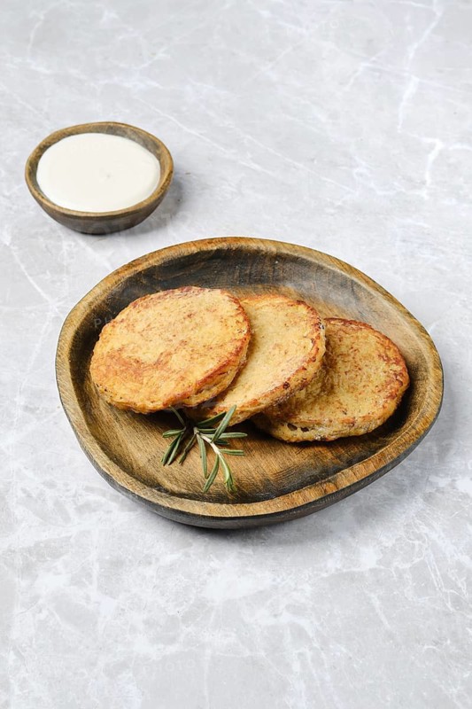 Golden potato cakes on a rustic wooden plate, garnished with fresh rosemary. A small bowl of sour cream accompanies the dish, perfect for a savory snack