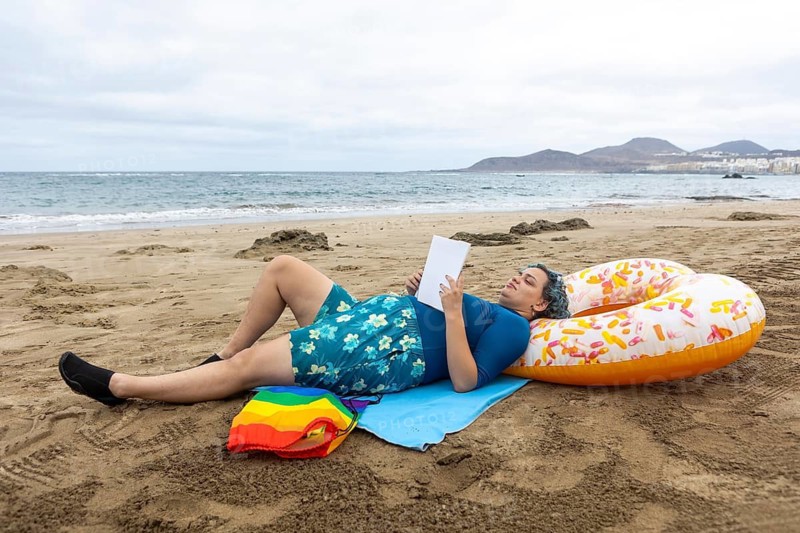 A transgender man lies on the sandy shore whilst engaging in a book, casually propped against a colorful float, against a scenic ocean backdrop
