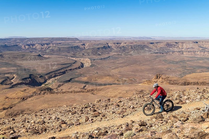 A man rides his mountain bike, fatbike, on the edge of the abyss, behind him the canyon landscape of the Fish River Canyon, Namibia