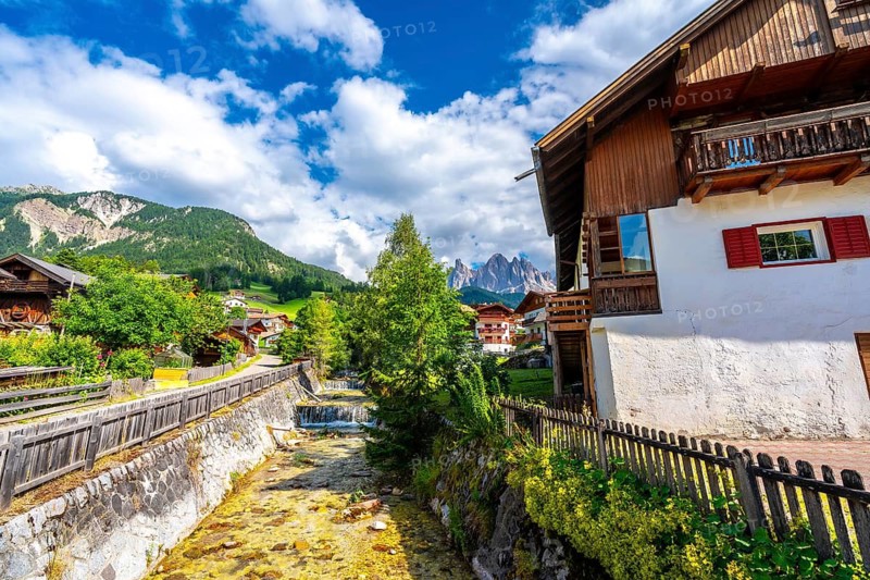 Scenic view of santa maddalena village and the odle mountains reflecting in a river during a sunny summer day in val di funes, dolomites, italy