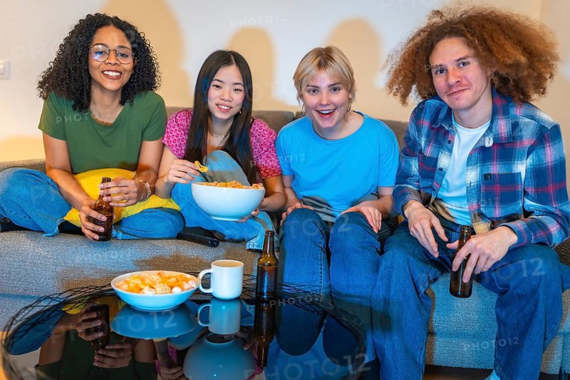 Four friends relaxing on a couch, sharing snacks and drinks while enjoying a movie together in a cozy living room setting