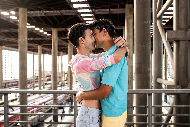 A joyful couple shares a loving embrace at a modern train station, their vibrant outfits enhancing the scene's warmth and vibrancy amidst the industrial setting of the station
