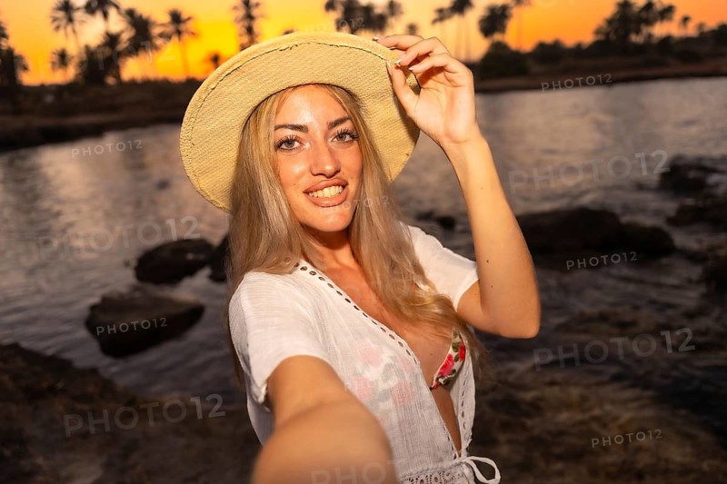 Personal point of view of a blonde beauty woman holding her hat while taking a selfie during sunset in a tropical beach
