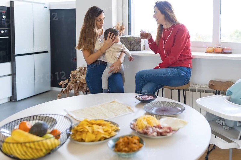Two mothers are chatting and drinking juice in a modern kitchen, preparing a meal of tacos and pizza with fresh ingredients, while holding a baby