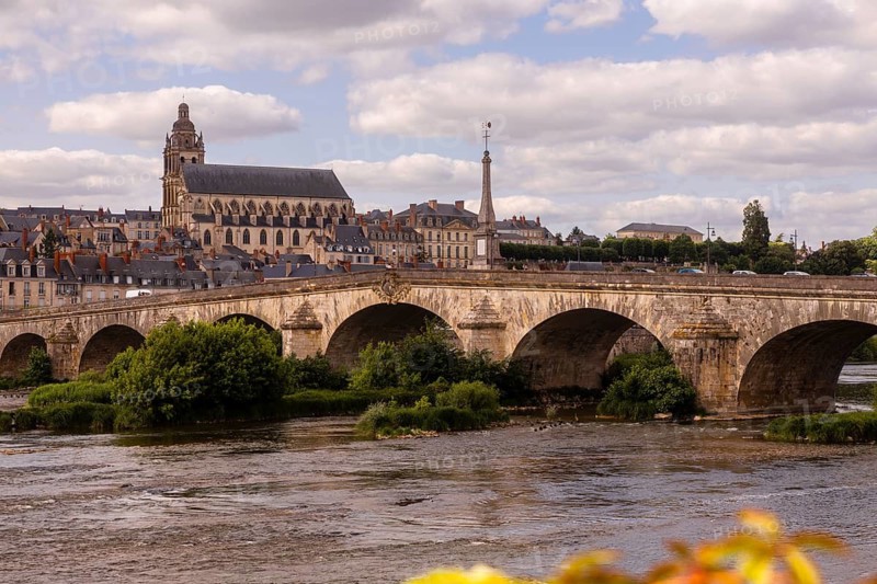 A picturesque view of Blois, France, showcasing its historic stone bridge and iconic skyline, featuring classic architecture under a partly cloudy sky. The river in the foreground adds tranquility