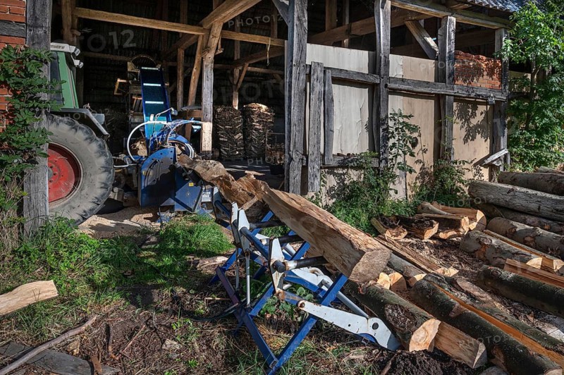 Young man sawing and splitting beech logs with his saw cutting machine, powered by a tractor in a barn, Othenstorf, Meckleburg-Vorpommern, Germany