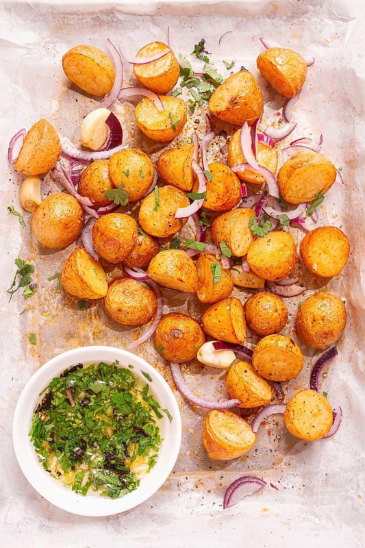 Roasted potatoes sit on a baking sheet next to chopped herbs and red onions. They are golden brown and ready to be served. Ingredients add color and flavor to the dish