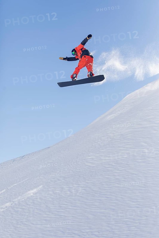 A snowboarder performs an impressive aerial trick, captured mid flight against a crisp blue sky. The snowy slope beneath adds to the dynamic, high adrenaline atmosphere