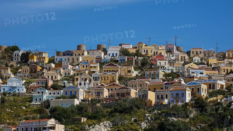 Colourful houses on a hill overlooking the sea under a bright blue sky, Symi Town, Symi, Dodecanese, Greek Islands, Greece