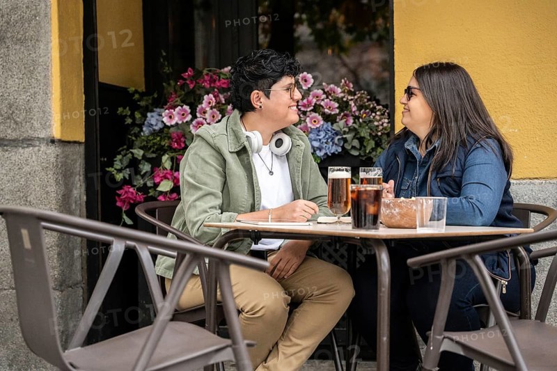A curvy lesbian couple shares a joyful moment at an outdoor beer. They are sitting at a table with drinks, surrounded by vibrant flowers, expressing happiness and affection