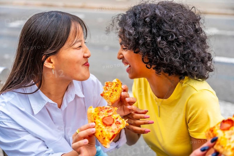 Multi-ethnic female friends smiling eating pizza sitting on steps in the street