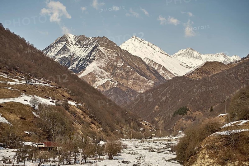 Majestic Caucasus snowy mountain peaks tower over a tranquil valley, where a rustic cabin nestled among bare trees creates a serene winter scene under a clear blue sky in the border with Georgia