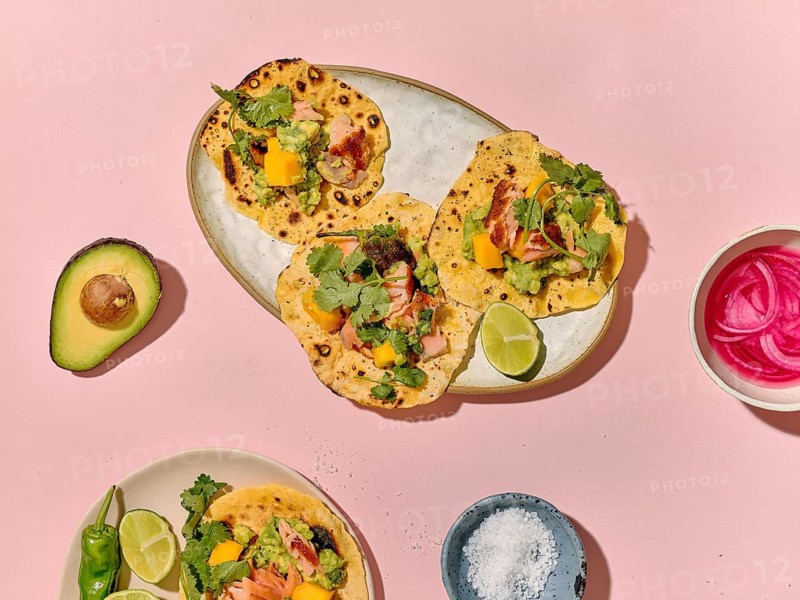Top view of colorful taco preparation showcasing fresh ingredients on a vivid pink background. Tacos with coriander, lime, avocado, and mango on plates are visible