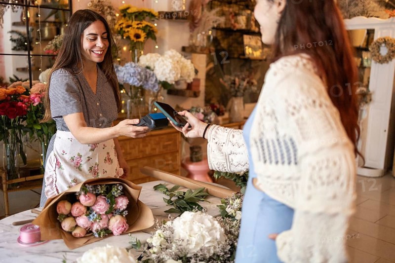 Two women are in a delightful florist shop, engaging in a friendly transaction. The shop is filled with beautiful flower arrangements, creating a lively and inviting atmosphere