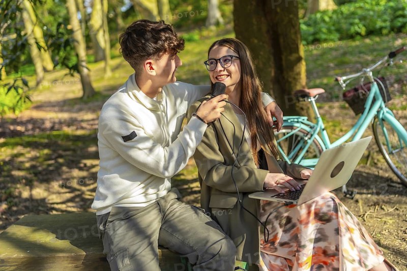 Two young journalists are recording a podcast in a park, using a microphone and laptop, with a bicycle in the background