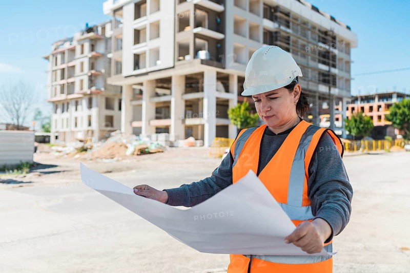 Woman in a hard hat and orange vest examines blueprints at a construction site. A modern building is visible in the background