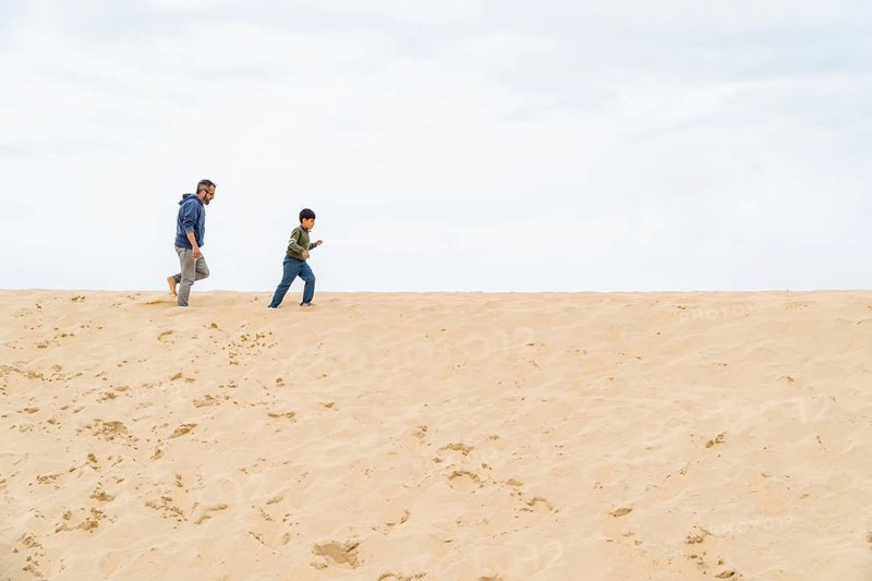 A father and son enjoy a carefree walk on a vast, sandy beach under a cloudy sky. The image captures a heartwarming moment of bonding and relaxation in the dune of Pilat