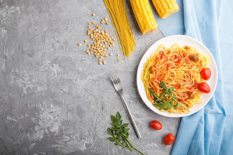 Corn noodles with tomato sauce and arugula on a gray concrete background and blue textile. Top view, flat lay, copy space