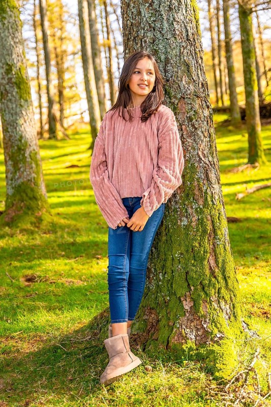 Vertical full length portrait of a cute teen girl leaning on tree trunk in an alpine forest