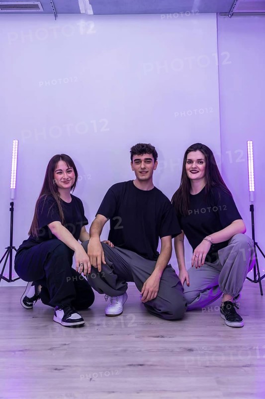 Three young dancers are crouching and posing together in a dance studio, illuminated by purple lights