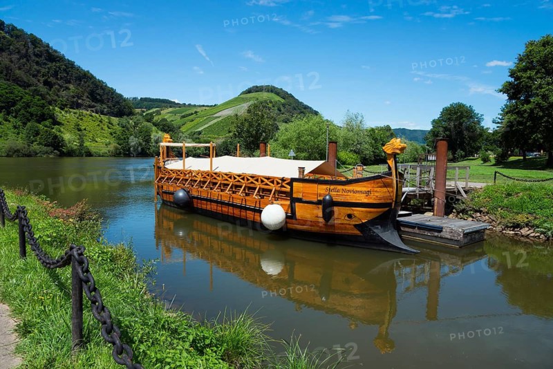 A wooden boat lies on a calm river in the middle of a green landscape with mountains and blue sky, Stella Noviomagi, wine ship monument, Neumagen wine ship, wooden replica of a Roman wine ship, Neumagen, Moselle, Rhineland-Palatinate, Germany