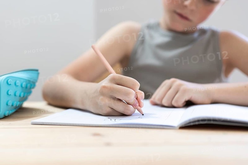 A young student concentrates on writing in a notebook with a pencil, a pencil case resting on the table. The scene captures a moment of focus and learning