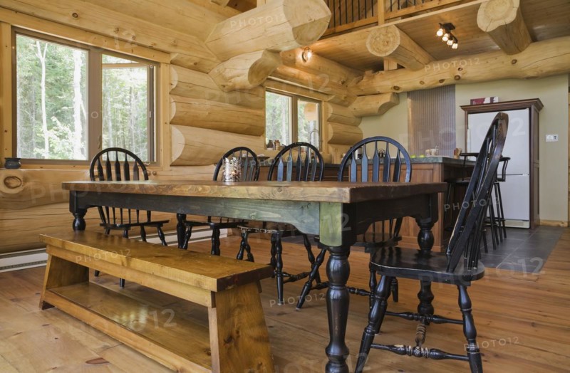 Black stained antique wood dining table with rounded high back chairs and pine wood sitting bench in open concept dining room and kitchen inside handcrafted Eastern white pine Scandinavian log cabin home, Quebec, Canada