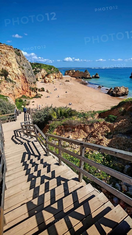 A wooden path, wooden staircase leads to a sandy beach with rocks and visitors, the still sea under a clear sky, Praia Dona Ana, Lagos, Algarve, Portugal