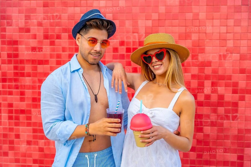 Young fashionable couple wearing summer clothes and sunglasses holding colorful drinks and smiling in front of a red tiled wall