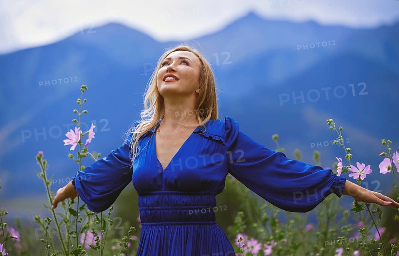 A joyful blond woman in a blue dress enjoys a moment of freedom surrounded by a vibrant grassy field. Her arms spread wide, embracing the tranquility of nature