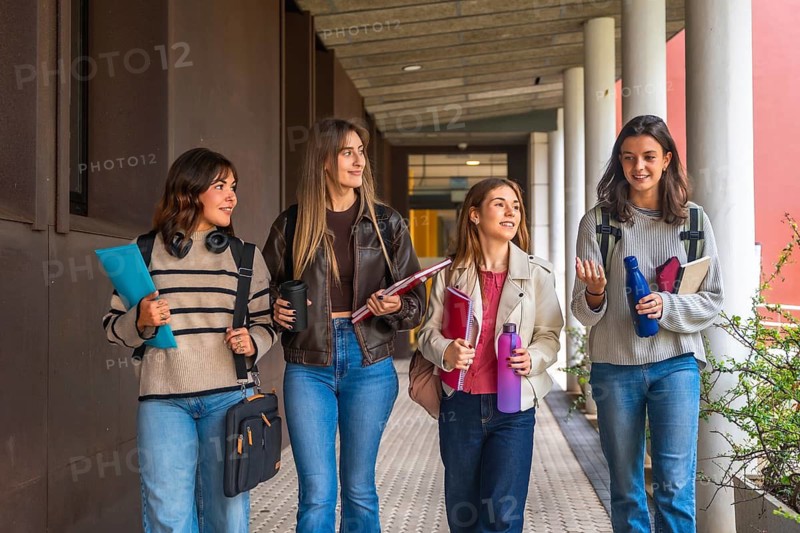 Four young women students are walking together along an outdoor university corridor, carrying books and bottles, engaging in conversation while heading to classes