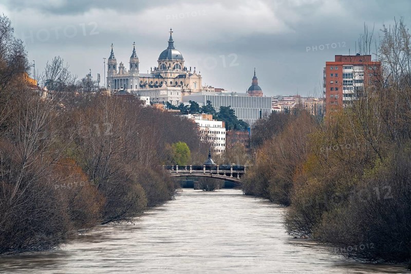 Captivating view of the Manzanares river flowing through Madrid, flanked by leafless trees. The cityscape in the background features the Almudena Cathedral under a cloudy sky