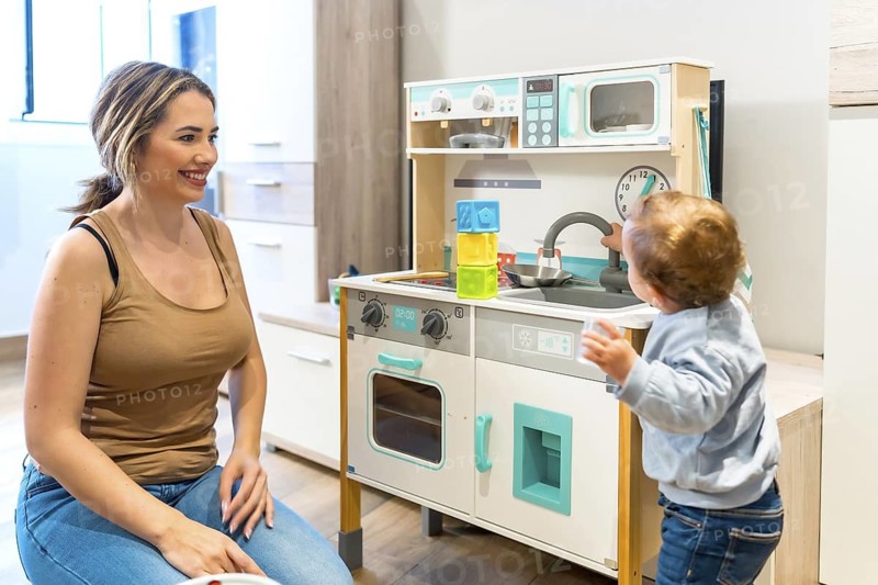 Happy mother watching her toddler son playing with a toy kitchen in a bright modern kitchen