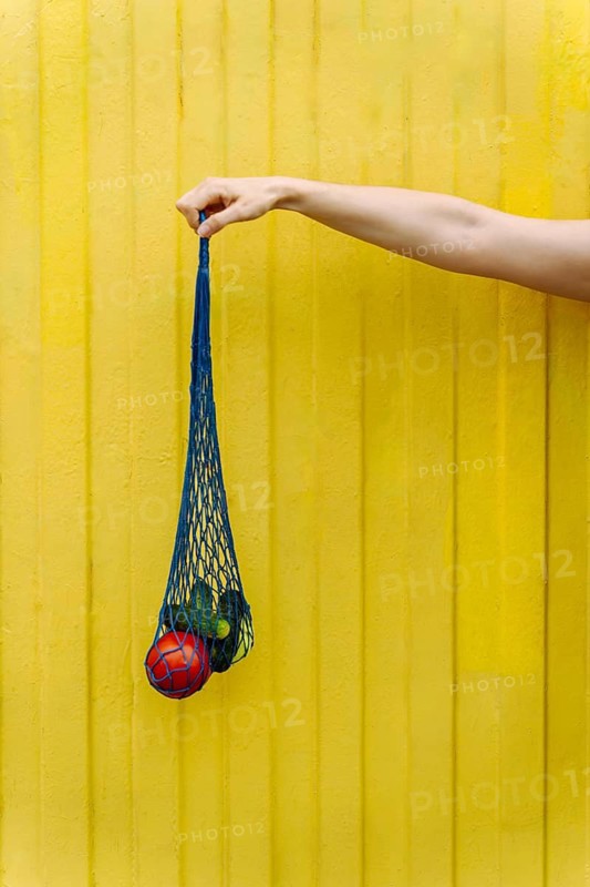 Hand holding mesh grocery bag with fresh tomatoes and cucumbers on yellow, A hand holds a blue mesh bag containing fruit and vegetables against a yellow background, conveying a minimalist and vibrant atmosphere