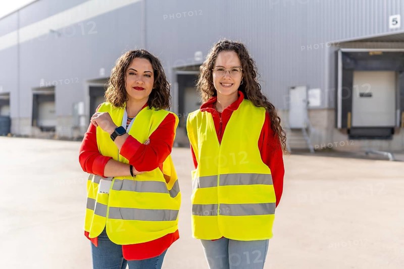 Portrait of distribution warehouse female colleagues standing in the parking lot smiling at camera wearing protective yellow reflective waistcoat