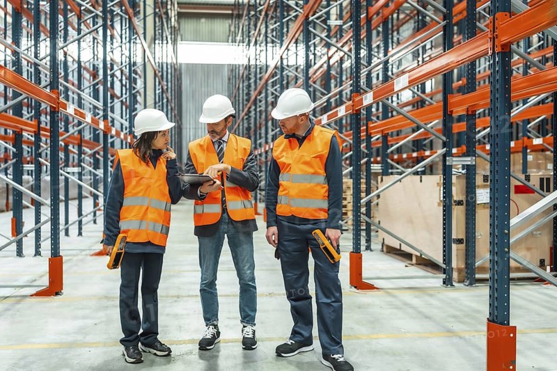 Warehouse workers wearing safety vests and hardhats are checking inventory using a digital tablet and barcode scanner in a large distribution center