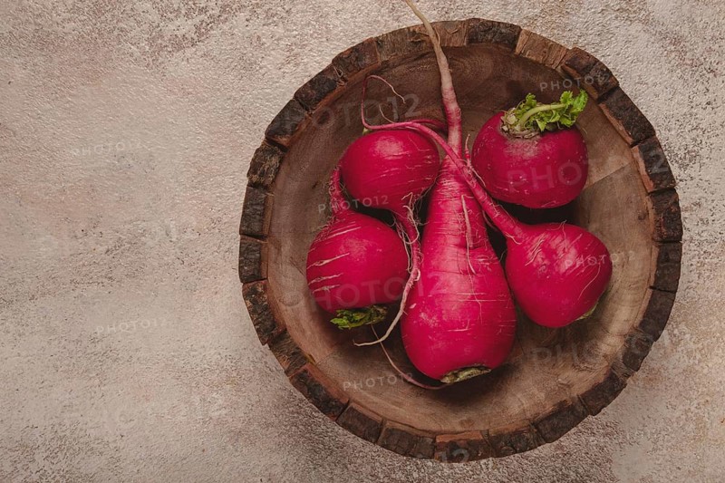 Vibrant red radishes with green tops arranged in a rustic wooden bowl on a textured surface create a visually appealing display. Perfect for highlighting fresh produce in a culinary setting