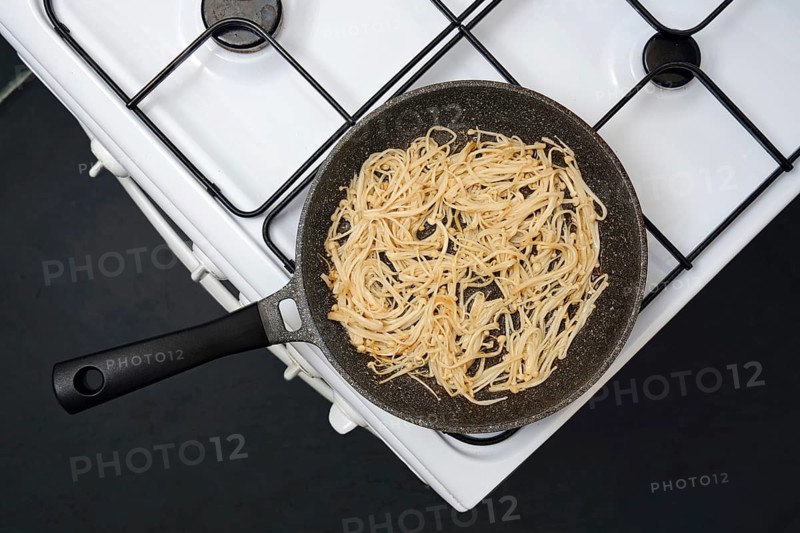 Overhead view of a frying pan with golden-brown enoki mushrooms on a gas stove, showing a homemade meal in the making. The kitchen setup highlights the cooking process.