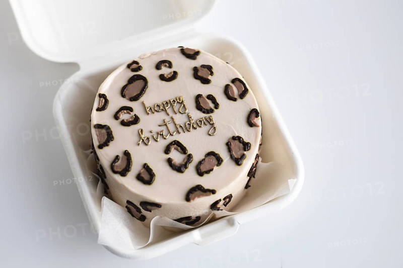 Top view of a stylish bento birthday cake with a leopard print pattern and a golden Happy Birthday message, served in a white takeout box
