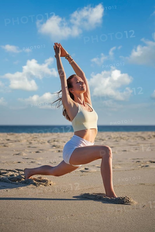 A woman performs Warrior 1 pose on the beach during sunset. She stretches her arms overhead while grounding her front foot. The calm ocean waves and picturesque sky create a serene backdrop