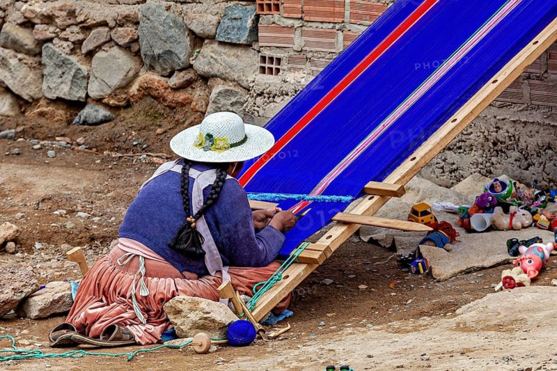 A woman in traditional garb carefully weaves on a blue loom in front of a brick wall, a woman weaves a cloth at Potosi on Cerro Rico in Bolivia