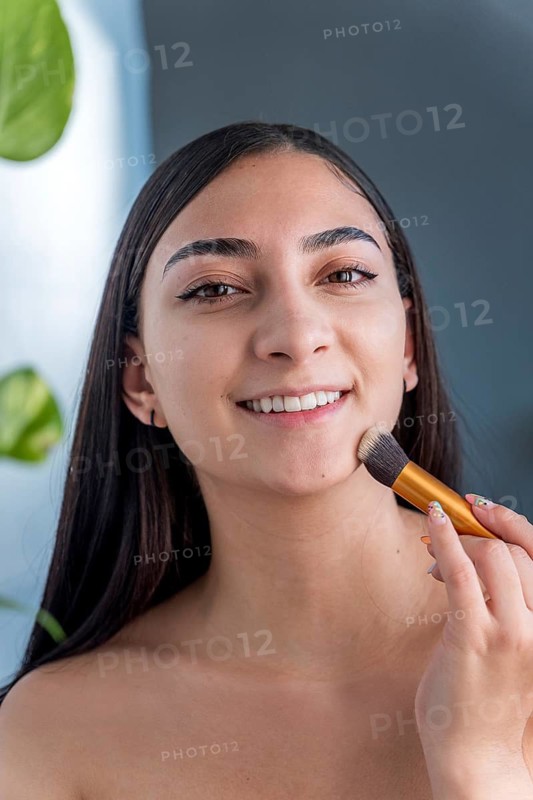 Smiling woman applying makeup with a brush, sitting in soft natural light. A tranquil scene highlighting beauty and self care, surrounded by a touch of greenery
