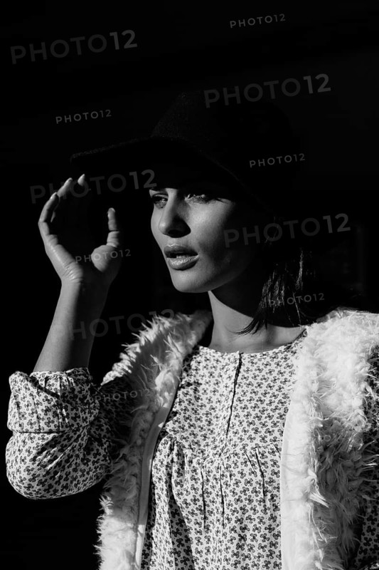 A striking black and white portrait of a woman wearing a hat and patterned outfit. The dramatic lighting highlights her elegant features and stylish attire