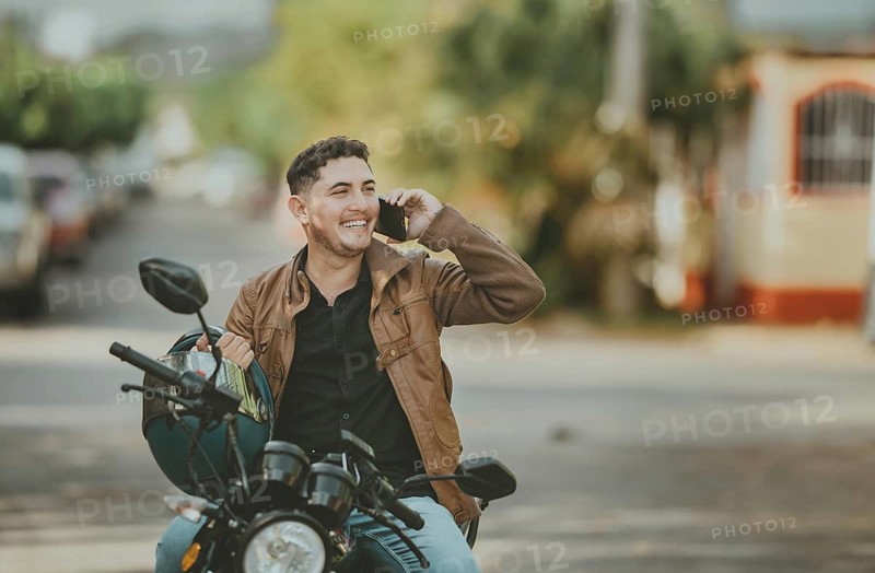 Handsome motorcyclist talking on the phone, with copy space. Smiling young motorcyclist in jacket talking by phone outdoors