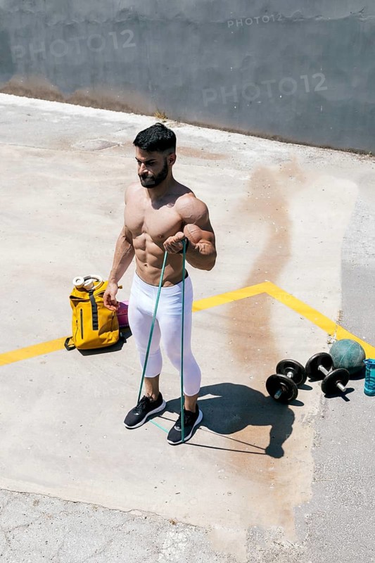 A fit man performs a bicep curl using a resistance band outdoors on a sunny day. He stands confidently on pavement, surrounded by workout equipment including dumbbells and a water bottle